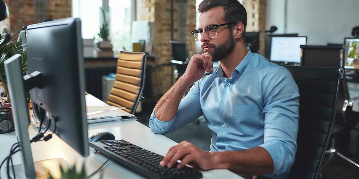 Seitenansicht eines jungen, bärtigen Mannes mit Brille und formeller Kleidung, der am Computer arbeitet und sein Kinn berührt, während er in einem modernen Büro sitzt