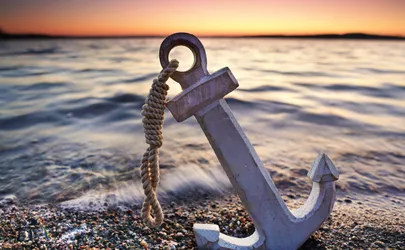 Ein Anker liegt am Strand; im Hintergrund das Meer und der Sonnenuntergang