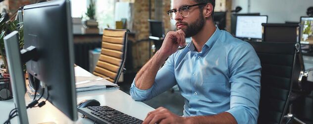 Seitenansicht eines jungen, bärtigen Mannes mit Brille und formeller Kleidung, der am Computer arbeitet und sein Kinn berührt, während er in einem modernen Büro sitzt