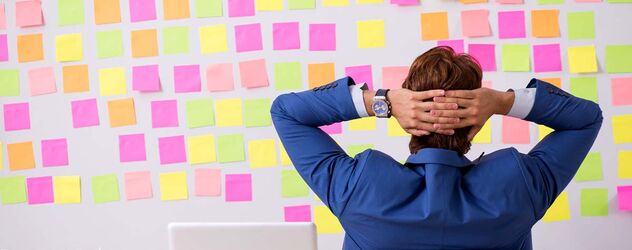ein Mann in Anzug hält seine Hände überkreuzt an seinem Hinterkopf und starrt eine Wand voller Notizen an