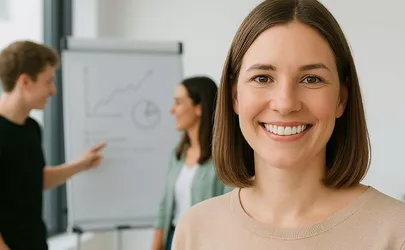eine Ausbilderin lächelt in der Kamera, zwei Azubis analysieren ein Projekt im Hintergrund am Flipchart