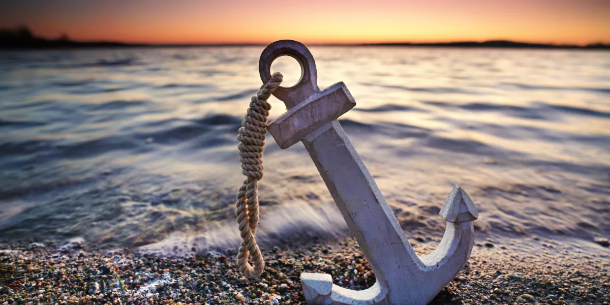 Ein Anker liegt am Strand; im Hintergrund das Meer und der Sonnenuntergang